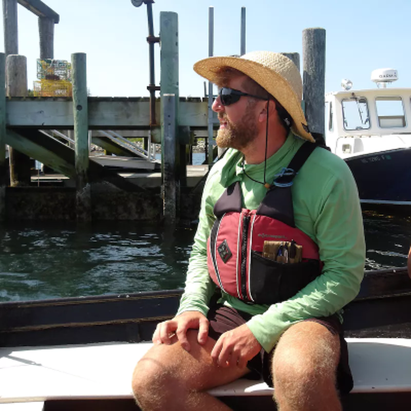 The image shows a man wearing a straw hat, sunglasses, and a life vest, sitting on a boat. He is looking to the side, with a pier and a boat in the background. The day appears to be sunny. The man seems to be enjoying the day out on the water.